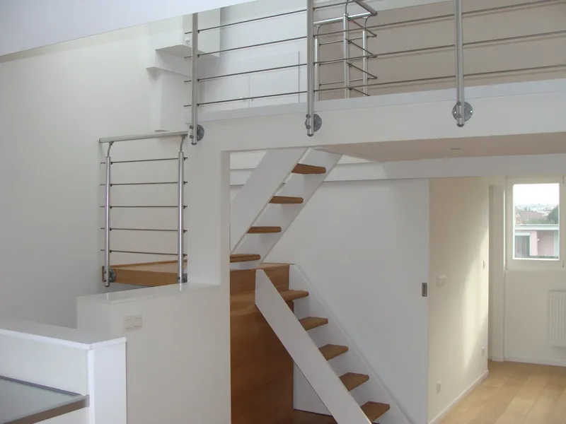 Escalier ouvert à marches en chêne et garde-corps en acier inoxydable, avec vide et mezzanine au-dessus.