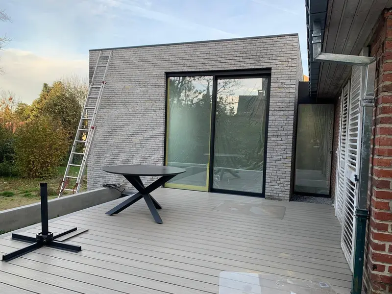 L’extension achevée en brique moulée main grise en lumière du soir, la maison existante et ses volets blancs à droite.